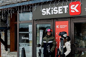 Two skiers standing with their gear outside a Skiset ski rental shop in Belle Plagne.