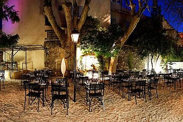 image of a courtyard at night-time with large trees, gravel floor and black chairs and tables