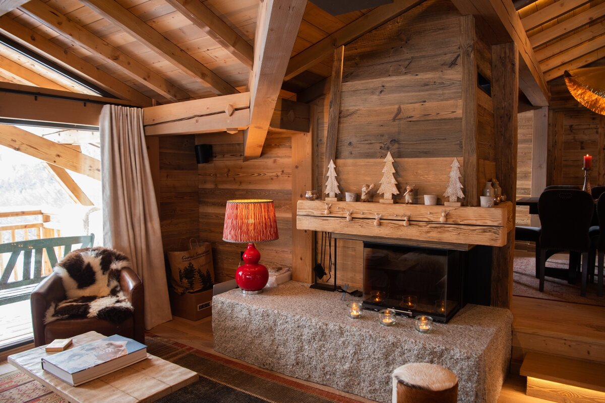 A cozy, wood-clad chalet living room features a stone fireplace with festive decor, a red lamp, armchair, and a large window overlooking outside.