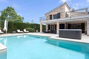 A luxurious stone house with a large swimming pool, hot tub, and outdoor seating under a clear blue sky. Lush greenery surrounds the property.