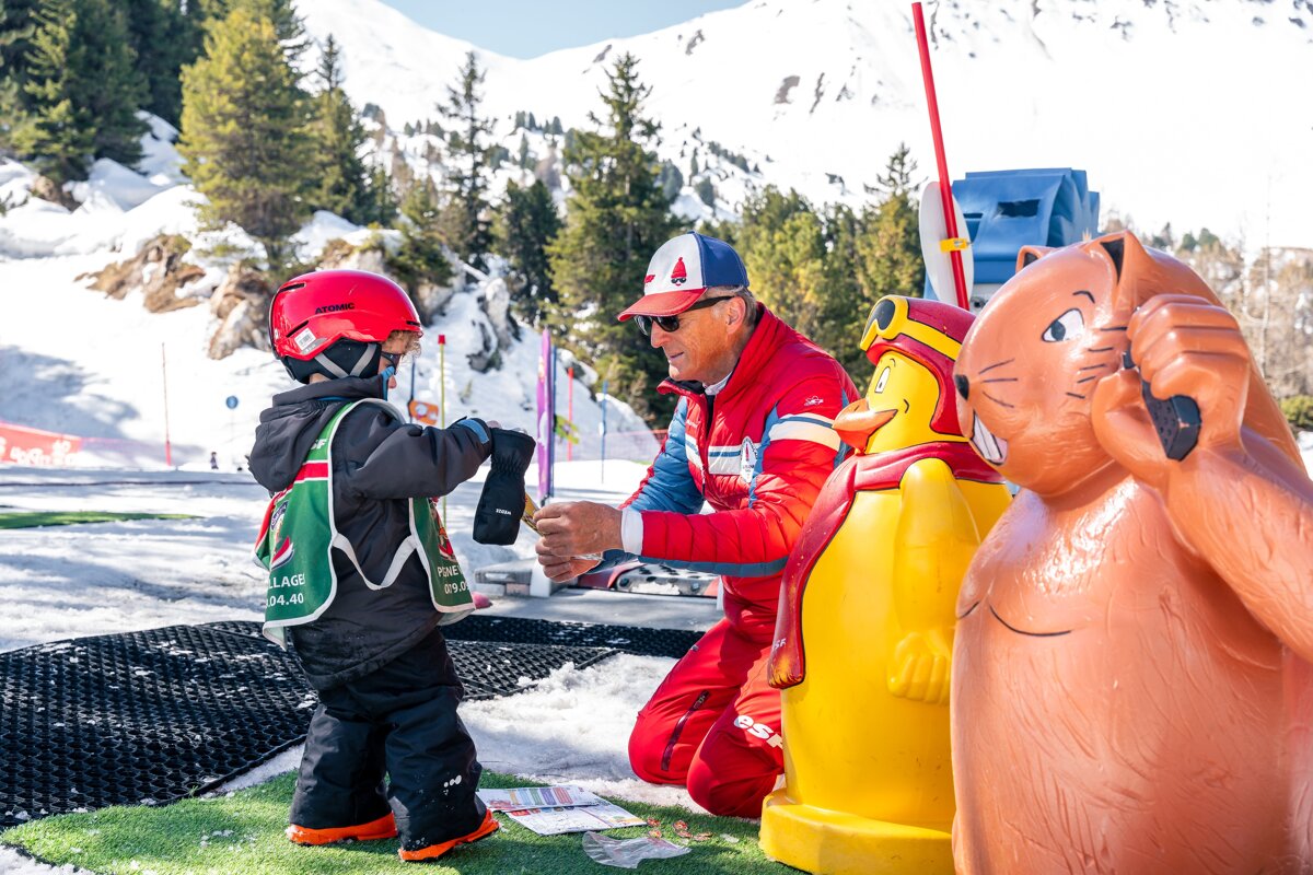 A ski instructor assists a child in a red helmet on a snowy mountain slope, with colorful cartoon figures beside them.