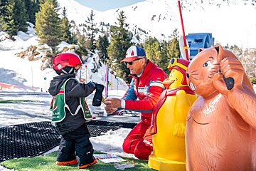 A ski instructor helps a young child in a red helmet with their glove at a kids' ski area, surrounded by snowy mountains and cartoon characters.