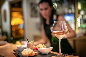A woman is standing behind a bar with a glass of wine and a plate of food