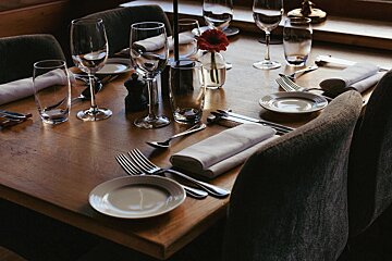 A beautifully set wooden dining table with elegant place settings, including various glasses, polished cutlery, white napkins, and a single red flower.