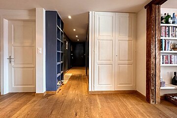 A warm-toned wooden floor leads through a hallway with blue shelves, white cabinets, and a rustic wooden beam beside a built-in bookshelf.