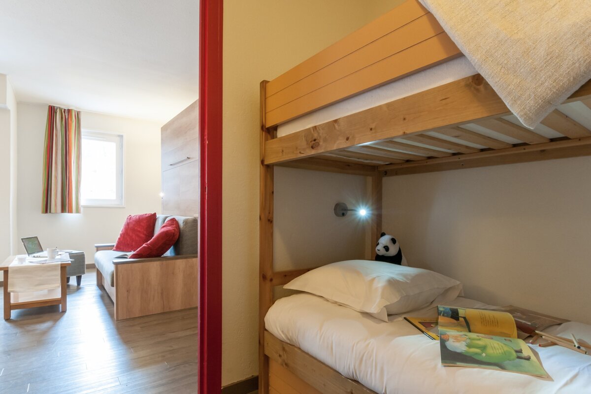 A cozy room featuring a wooden bunk bed in the foreground and a view into a living area with a sofa and coffee table.