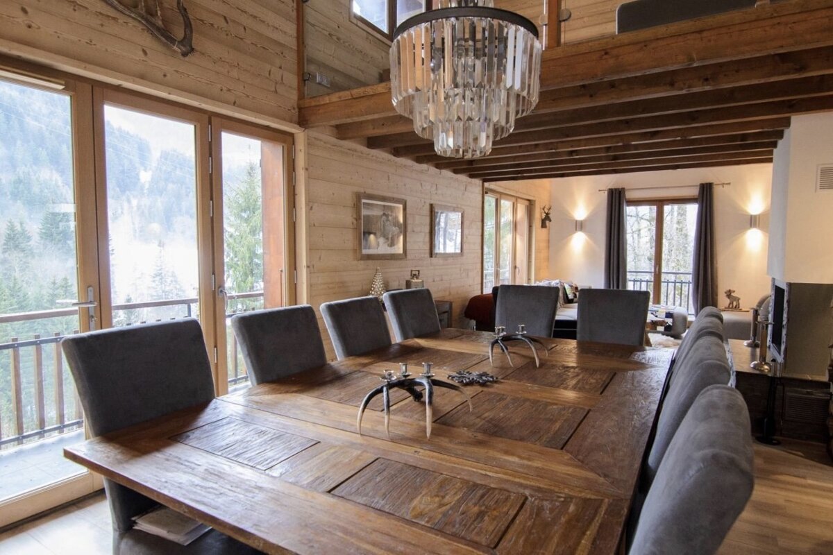 A rustic wooden dining room features a large table with grey chairs, a crystal chandelier, and expansive windows offering a mountain forest view.