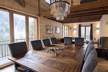 A rustic wooden dining room features a large table with grey chairs, a crystal chandelier, and expansive windows offering a mountain forest view.