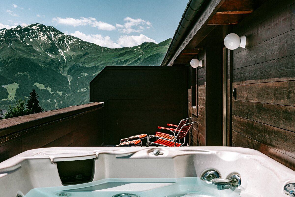 A luxurious hot tub on a wooden balcony offers stunning views of green, snow-capped mountains under a blue sky with clouds. Red chairs are visible on the deck.