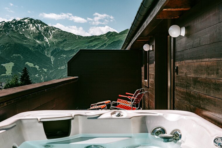 A luxurious hot tub on a wooden balcony offers stunning views of green, snow-capped mountains under a blue sky with clouds. Red chairs are visible on the deck.