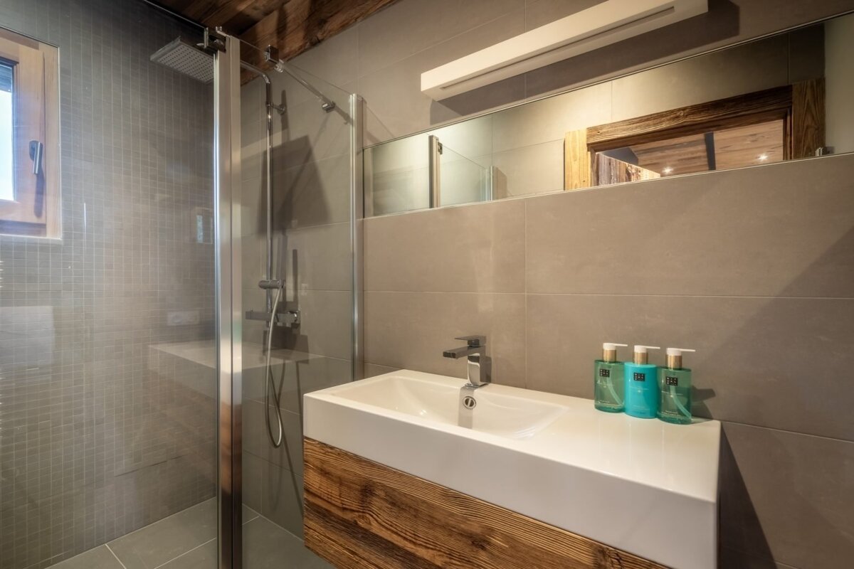 A contemporary bathroom with grey tiled walls, glass shower, wooden vanity, and white sink. Exposed wood beams and a mirror complete the modern look.