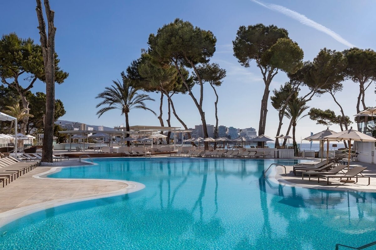 Sunny resort pool with turquoise water, surrounded by pine and palm trees, lounge chairs, umbrellas, and distant coastal buildings.