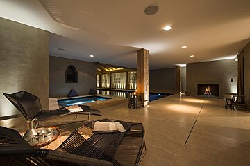 A luxurious indoor spa area featuring warm lighting, two pools, comfortable loungers, a cozy fireplace, and rustic wooden accents, perfect for relaxation.