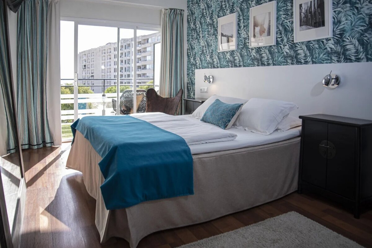 Bright hotel room features a bed with blue accents, palm leaf wallpaper, and a balcony overlooking city buildings.