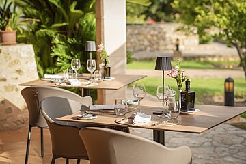 Tables set for outdoor dining at a restaurant, featuring wine glasses, small lamps, and an olive oil bottle, surrounded by lush greenery.