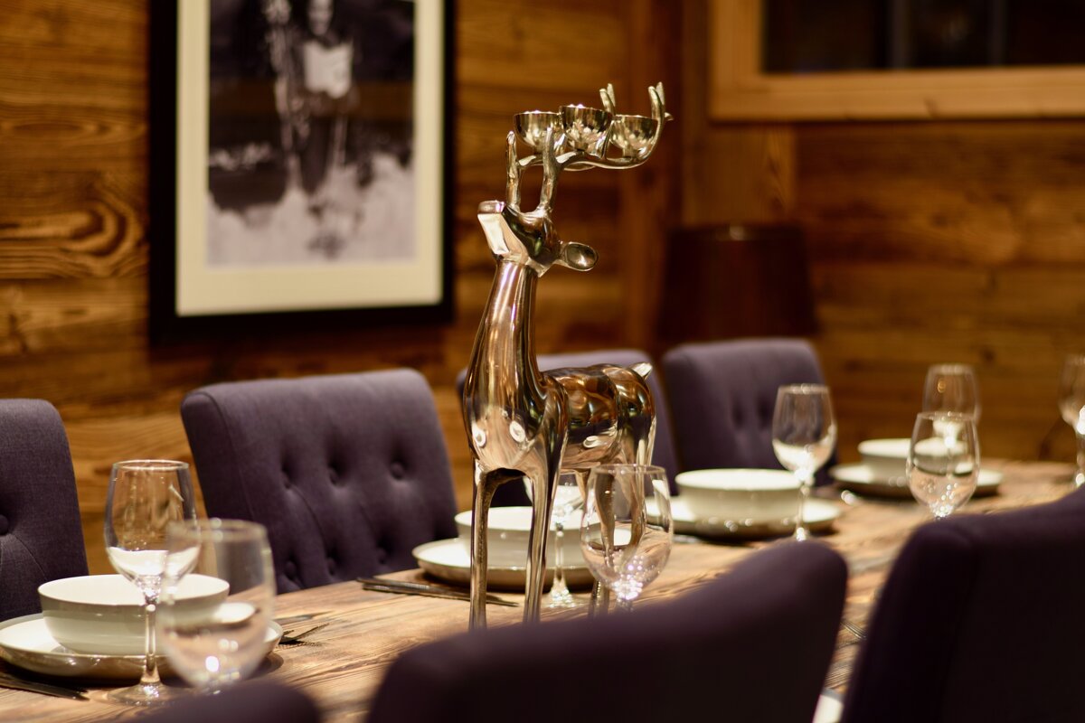 A rustic dining room features a wooden table set with plates, glasses, and purple chairs. A prominent silver deer candlestick holder serves as the centerpiece.
