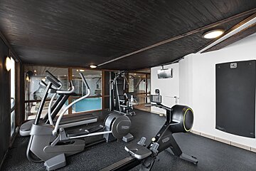 An indoor gym featuring an elliptical, a rowing machine, and strength equipment under a dark wooden ceiling.