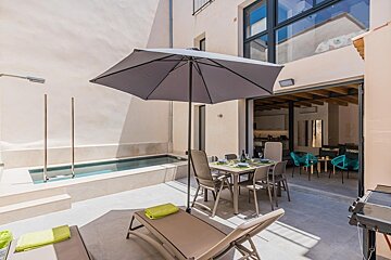 A sunny modern patio features a small pool, umbrella-shaded dining table, sun loungers, and an open-plan connection to an indoor living area.