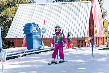 A smiling child in a pink ski suit, helmet, and goggles stands on skis at a sunny ski school, with buildings and a lift behind.