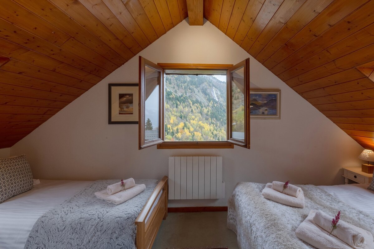 A cozy attic bedroom with two twin beds and a rustic wooden ceiling. An open window offers a breathtaking view of a mountainside covered in autumn foliage.