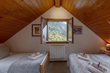 A cozy attic bedroom with two twin beds and a rustic wooden ceiling. An open window offers a breathtaking view of a mountainside covered in autumn foliage.
