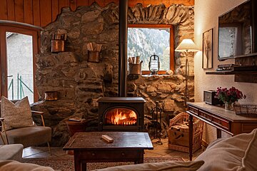 A cozy, rustic room features a blazing wood stove, stone walls, and a window revealing a mountain view. Warm lighting and comfortable seating create an inviting atmosphere.
