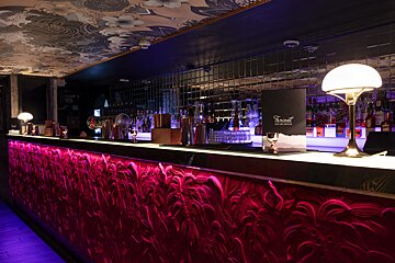A stylish bar featuring an illuminated magenta front with swirling patterns, a mirrored tiled backsplash, and a decorative patterned ceiling. The counter holds drinks, shakers, and elegant lamps.