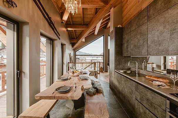 A modern chalet kitchen/dining area features a rustic live-edge wood table, sleek gray cabinetry, wooden beams, and large windows with snowy mountain views.