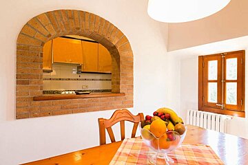 A bright dining space featuring a brick archway connecting to a kitchen. A bowl of fruit sits on a wooden table, with a wooden-framed window nearby.