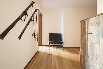 Bright hallway with wooden floor, vintage skis and poles on a white wall, a black armchair, and a reclaimed wood panel.