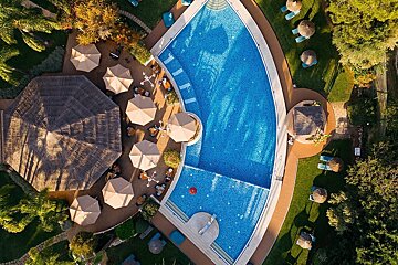 Aerial view of a vibrant blue curved resort swimming pool, surrounded by numerous beige umbrellas, loungers on green grass, and thatched buildings.