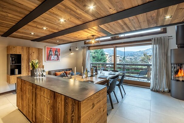 A cozy, modern open-plan kitchen and dining area with a wood ceiling, fireplace, and large window overlooking a scenic mountain view.