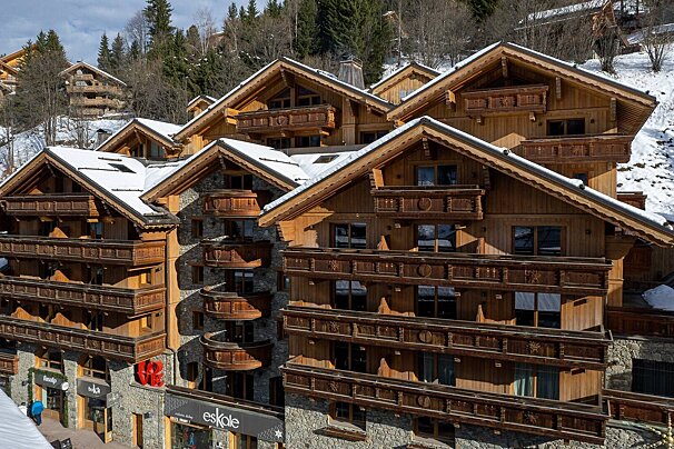Wooden chalets with snow-dusted roofs and ornate balconies ascend a sunny, tree-covered mountainside, showcasing classic alpine architecture.