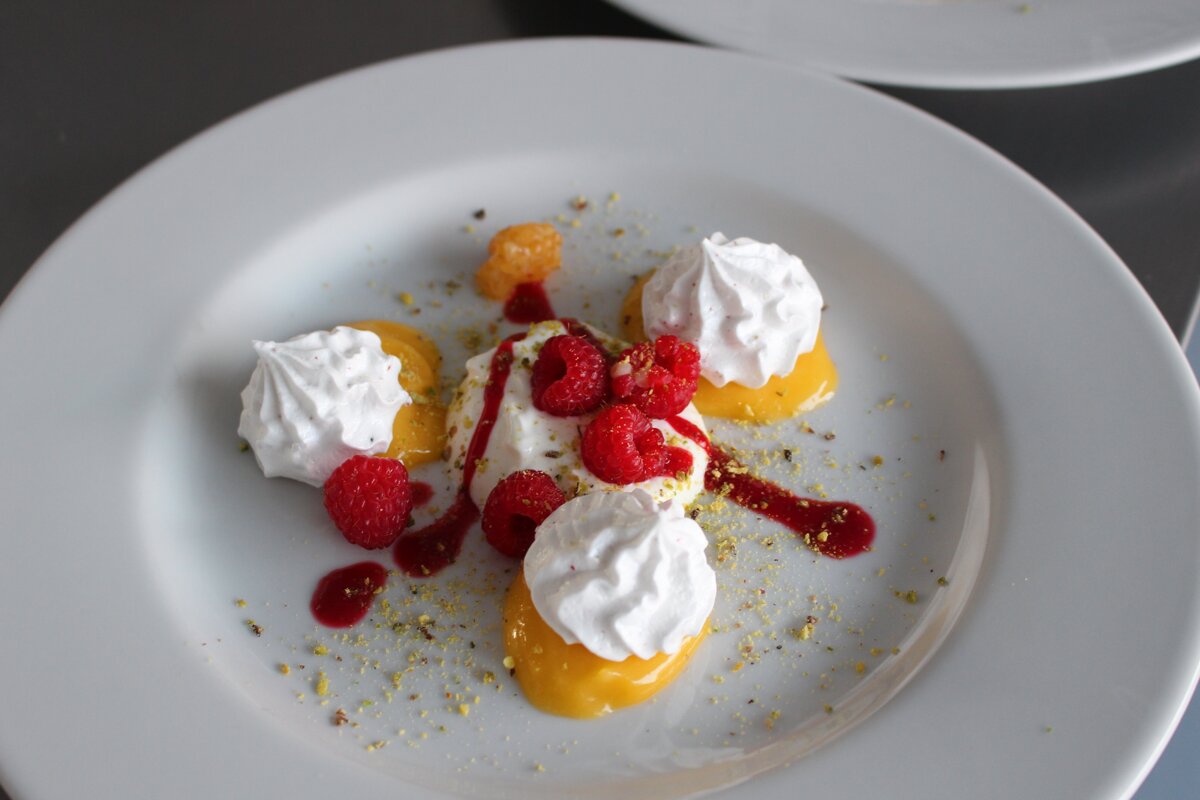 A gourmet dessert on a white plate, featuring mango puree, meringues, fresh raspberries, raspberry coulis, and pistachio dust.
