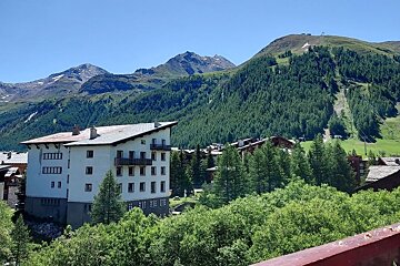 A white building with balconies amidst a lush green forest in a mountain village, set against a backdrop of towering mountains and a clear blue sky.