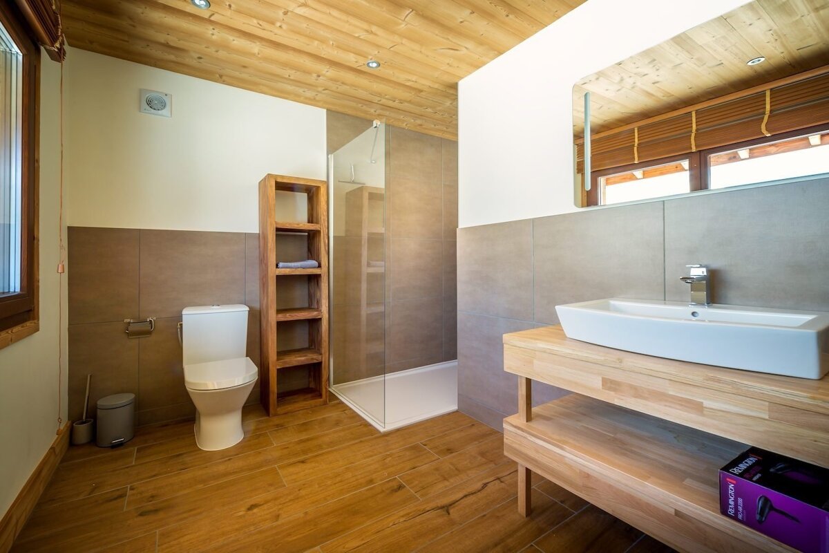 Modern bathroom featuring wooden floors/ceiling, white toilet, glass shower, and a white sink on a wooden vanity. Walls are white and grey tiled.
