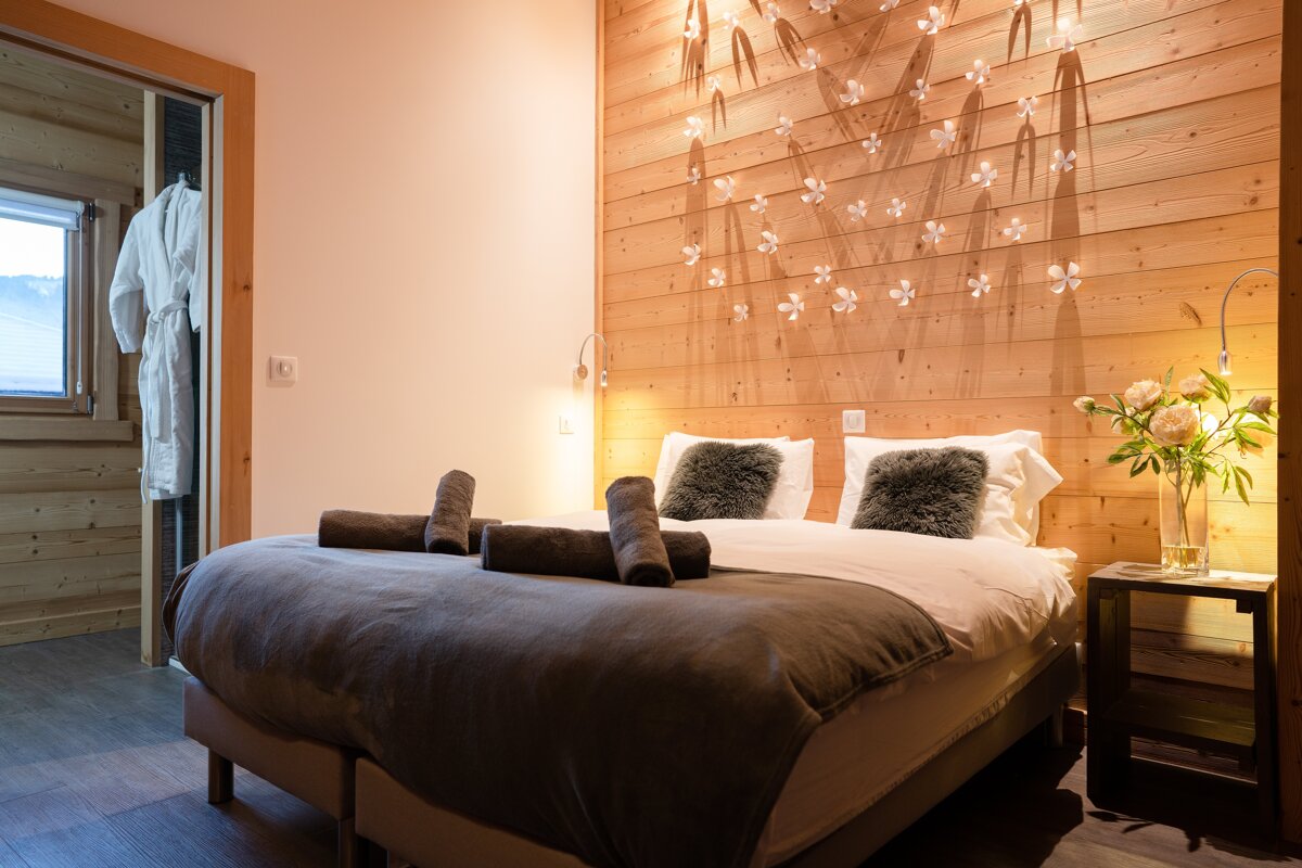 A warm, rustic bedroom with a wooden accent wall featuring delicate floral decor. A comfy bed with gray bedding and towels, and a white robe in the doorway.