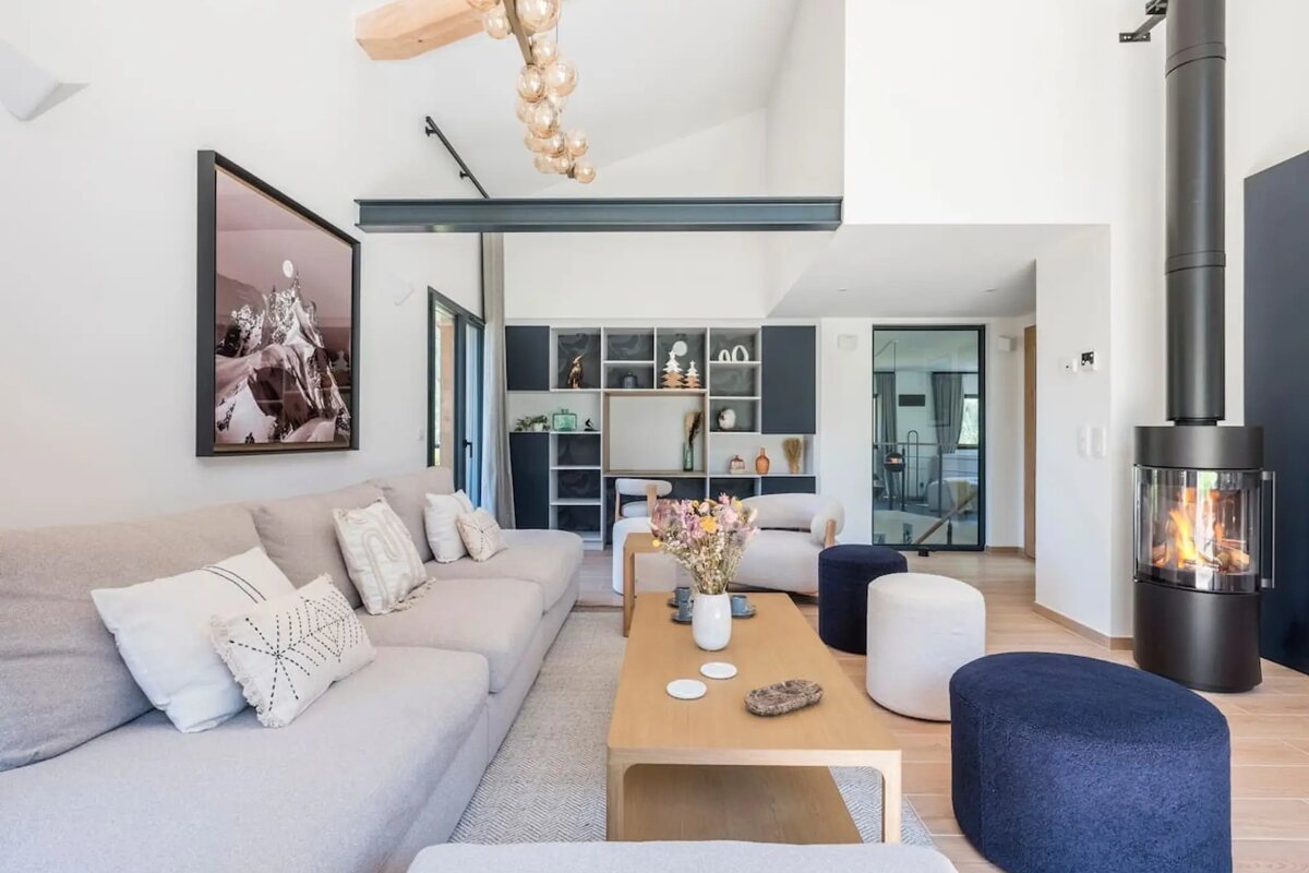 A chic, modern living room featuring a light grey sectional sofa, wooden coffee table, round ottomans, and a cozy, burning wood stove.