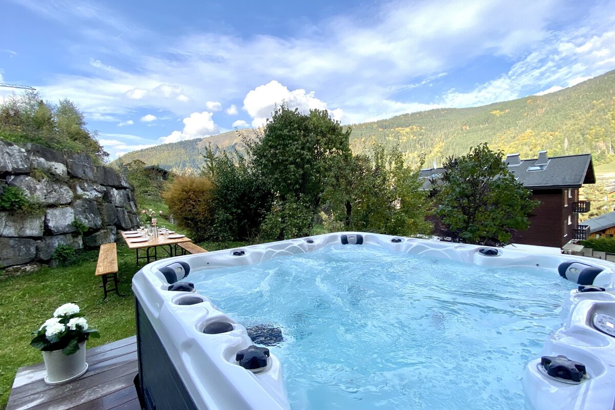 A bubbly outdoor hot tub and dining table with mountain views under a blue, cloudy sky. Green foliage and a stone wall complete the serene scene.