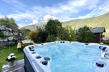 A bubbly outdoor hot tub and dining table with mountain views under a blue, cloudy sky. Green foliage and a stone wall complete the serene scene.