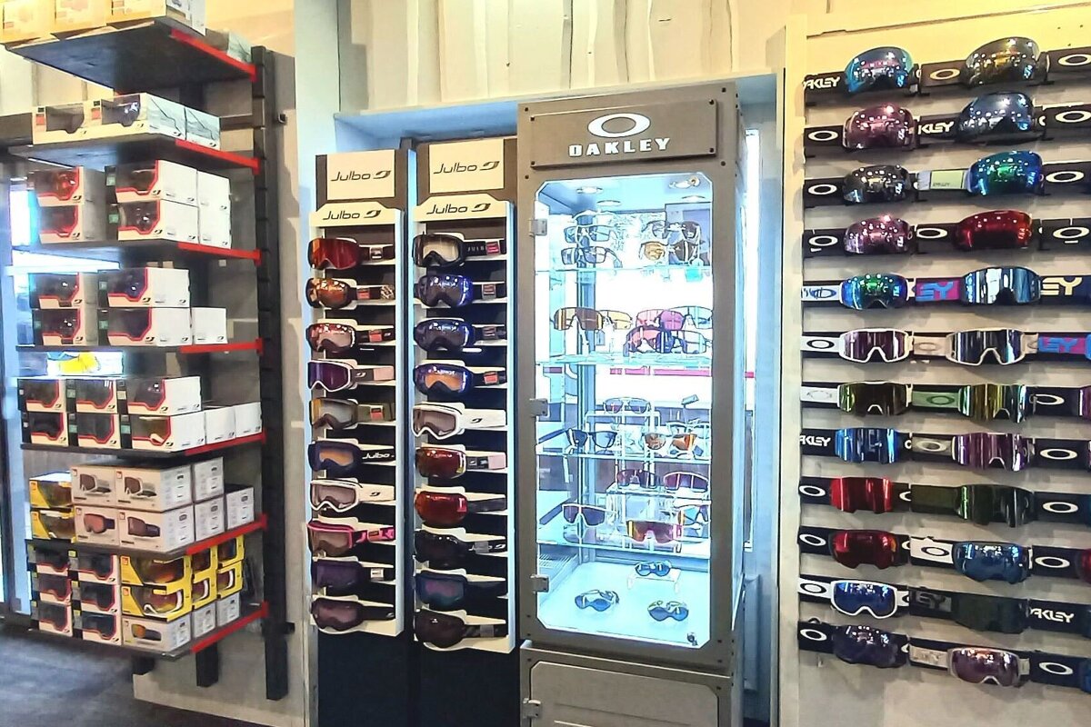 A store displays numerous colorful ski goggles from Julbo and Oakley, with Oakley sunglasses in a lit central cabinet and boxed stock on shelves.
