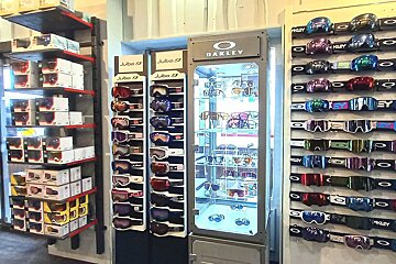 A store displays numerous colorful ski goggles from Julbo and Oakley, with Oakley sunglasses in a lit central cabinet and boxed stock on shelves.