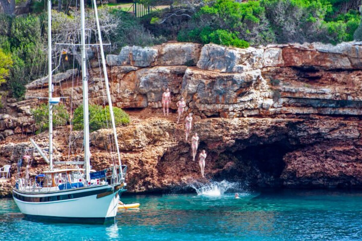 People are jumping off a cliff into the water near a sailboat