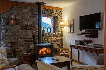 A cozy, rustic living room with a stone wall, burning wood stove, mountain view from the window, TV, and warm lighting.