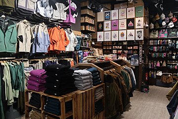 A bustling clothing store featuring racks of colorful jackets, folded apparel on wooden displays, and various accessories like hats and gloves. Posters decorate a wall.