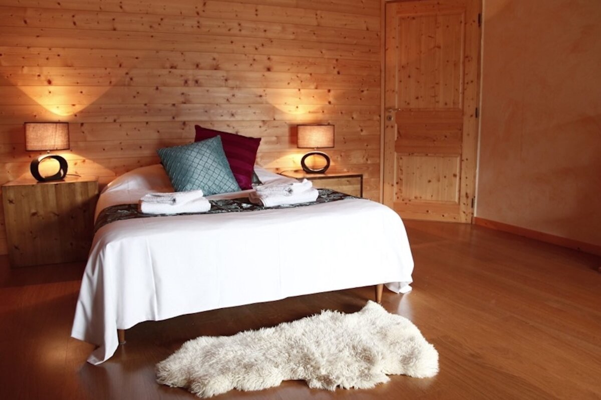 A cozy, wood-paneled bedroom features a white bed with colorful pillows, two bedside lamps, a wooden floor, and a fluffy white rug.