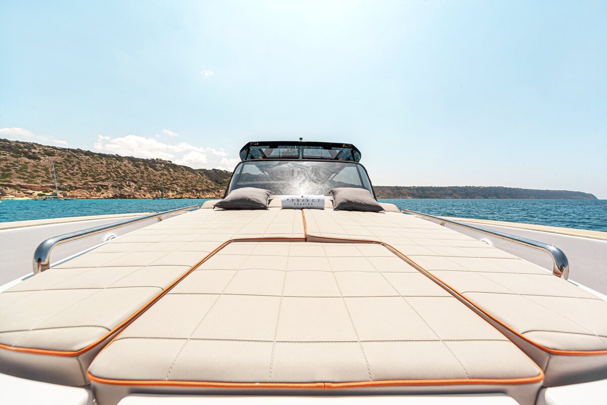 Front deck of a luxury yacht with beige sun pads and pillows, sailing on calm blue waters near a rocky coastline.