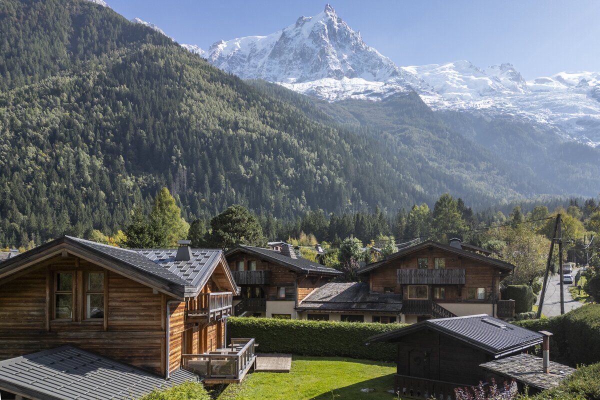 Charming wooden chalets in an alpine valley, backed by lush green forested mountains and majestic snow-capped peaks, including a prominent snowy pinnacle.