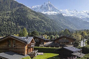 Charming wooden chalets in an alpine valley, backed by lush green forested mountains and majestic snow-capped peaks, including a prominent snowy pinnacle.