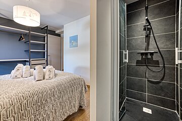 A modern bedroom with a bed, open closet, and an adjacent dark-tiled shower stall. White towels are neatly folded on the cozy bed.